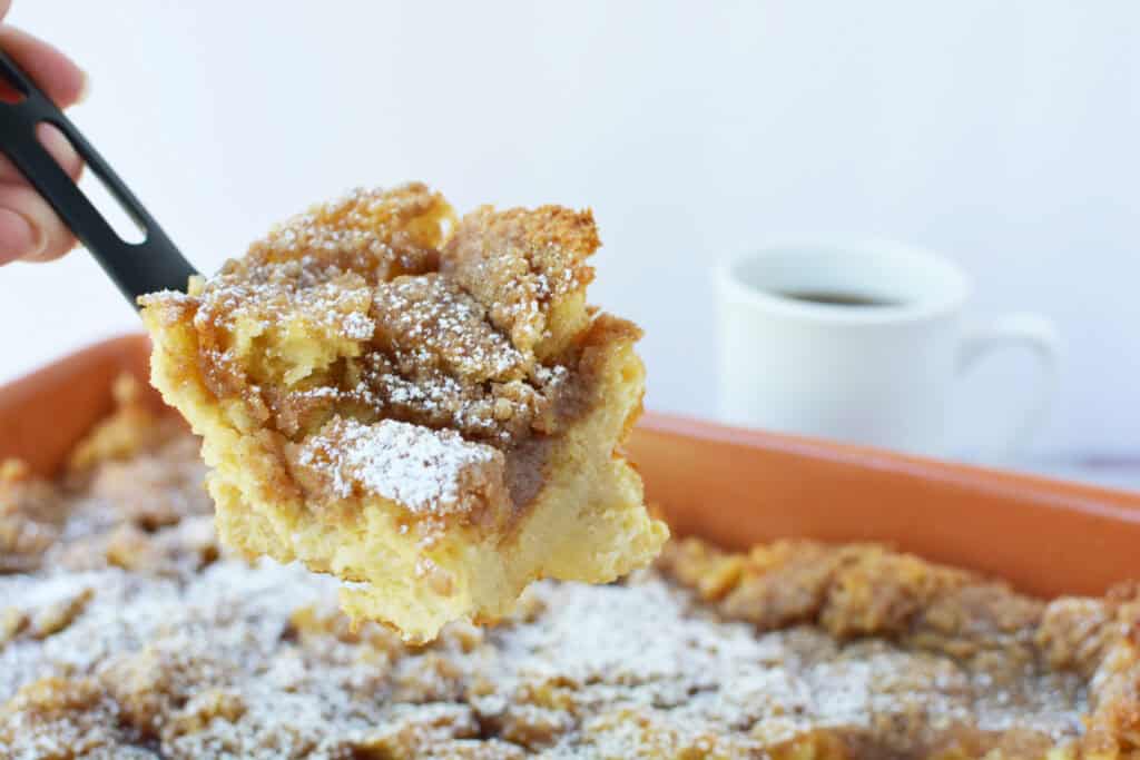 A spatula lifts a piece of Overnight French Toast Casserole topped with powdered sugar and crumb topping from a baking dish, with a white cup of coffee in the background.