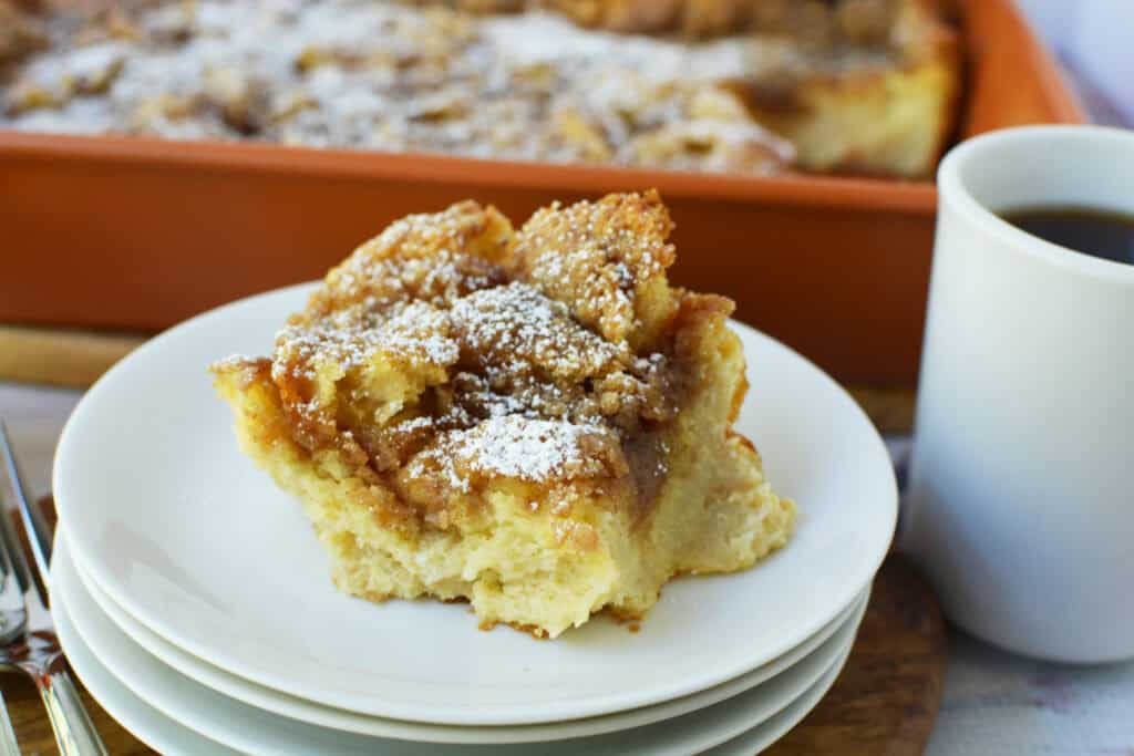 A slice of cinnamon coffee cake topped with powdered sugar sits on a stack of white plates beside a cup of black coffee, with the rest of the Overnight French Toast Casserole in a baking dish in the background.