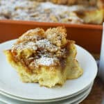 A slice of cinnamon coffee cake topped with powdered sugar sits on a stack of white plates beside a cup of black coffee, with the rest of the Overnight French Toast Casserole in a baking dish in the background.