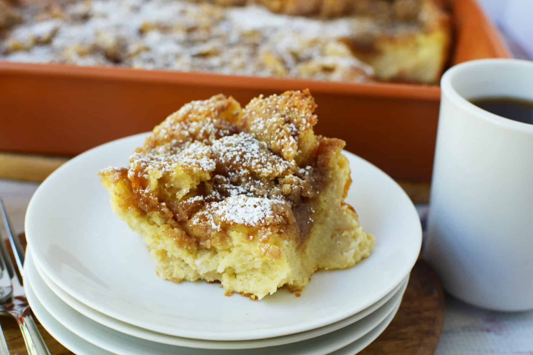 A slice of cinnamon coffee cake topped with powdered sugar sits on a stack of white plates beside a cup of black coffee, with the rest of the Overnight French Toast Casserole in a baking dish in the background.