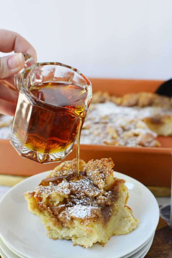 A hand pours syrup from a small glass pitcher onto a serving of Overnight French Toast Casserole topped with powdered sugar, placed on a white plate with a baking dish in the background.