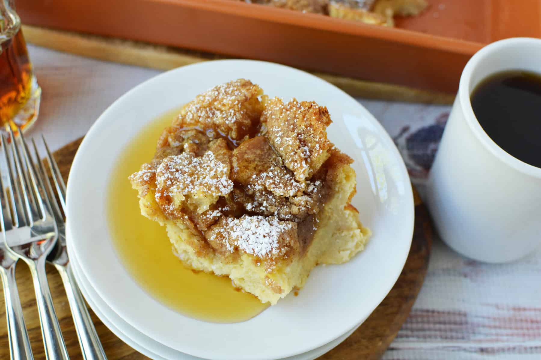 A slice of Overnight French Toast Casserole dusted with powdered sugar sits on a white plate with syrup, next to a cup of coffee and several forks, with the casserole dish and syrup bottle in the background.