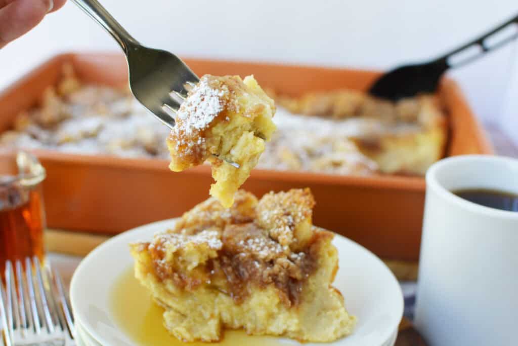 A fork lifts a bite of bread pudding, reminiscent of Overnight French Toast Casserole, topped with powdered sugar from a plate; a mug of coffee, syrup, and a baking dish with more bread pudding are in the background.