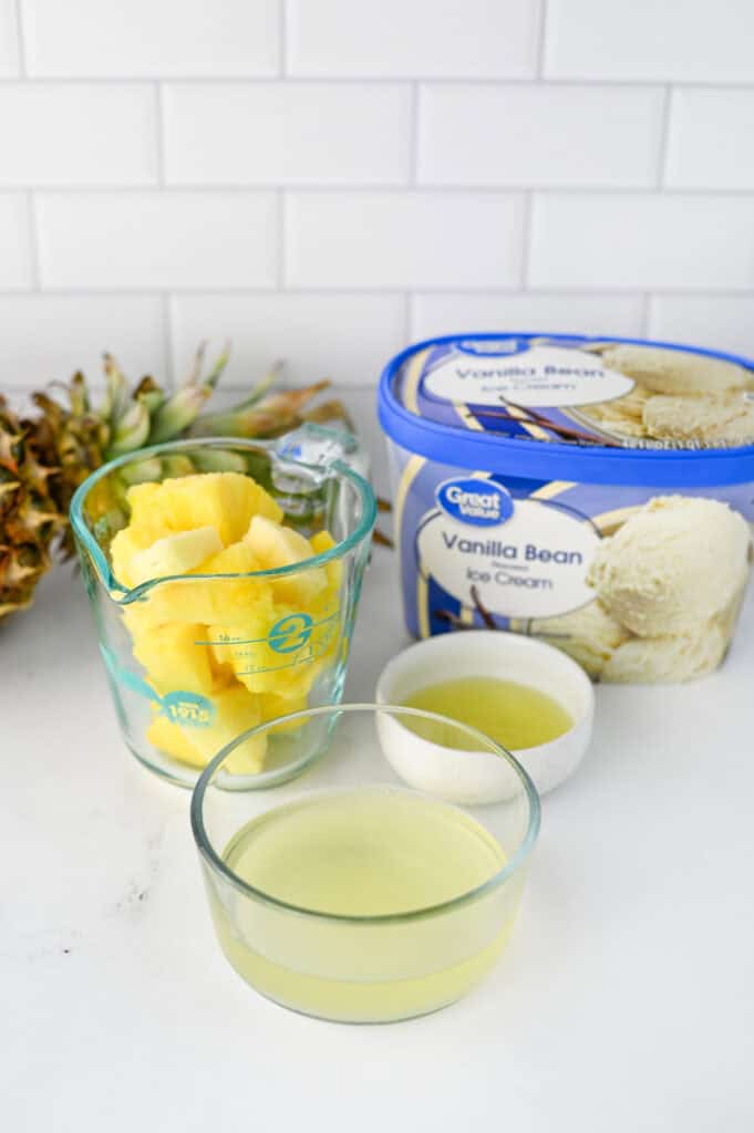 A measuring cup of fresh pineapple chunks, a bowl and a measuring cup of pineapple juice, a small bowl of lemon juice, and vanilla bean ice cream are arranged on a white counter—perfect ingredients for the classic Disney Dole Whip Recipe.