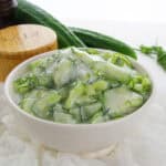 Wide view of cucumber salad in a white bowl with dill and a salt container nearby.