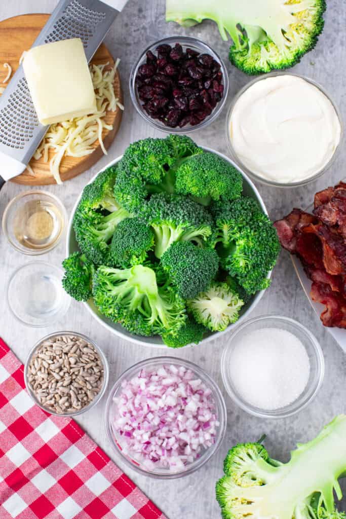 Overhead view of a costco broccoli salad in a bowl, surrounded by small bowls of shredded cheese, dried cranberries, sour cream, chopped red onion, sunflower seeds, sugar, vinegar, and cooked bacon on a gray surface with a red checkered napkin.