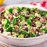 A large white bowl filled with a fresh Costco broccoli salad, featuring chopped broccoli, bacon bits, dried cranberries, sunflower seeds, and a creamy dressing. A red-checked napkin and lemons are in the background.