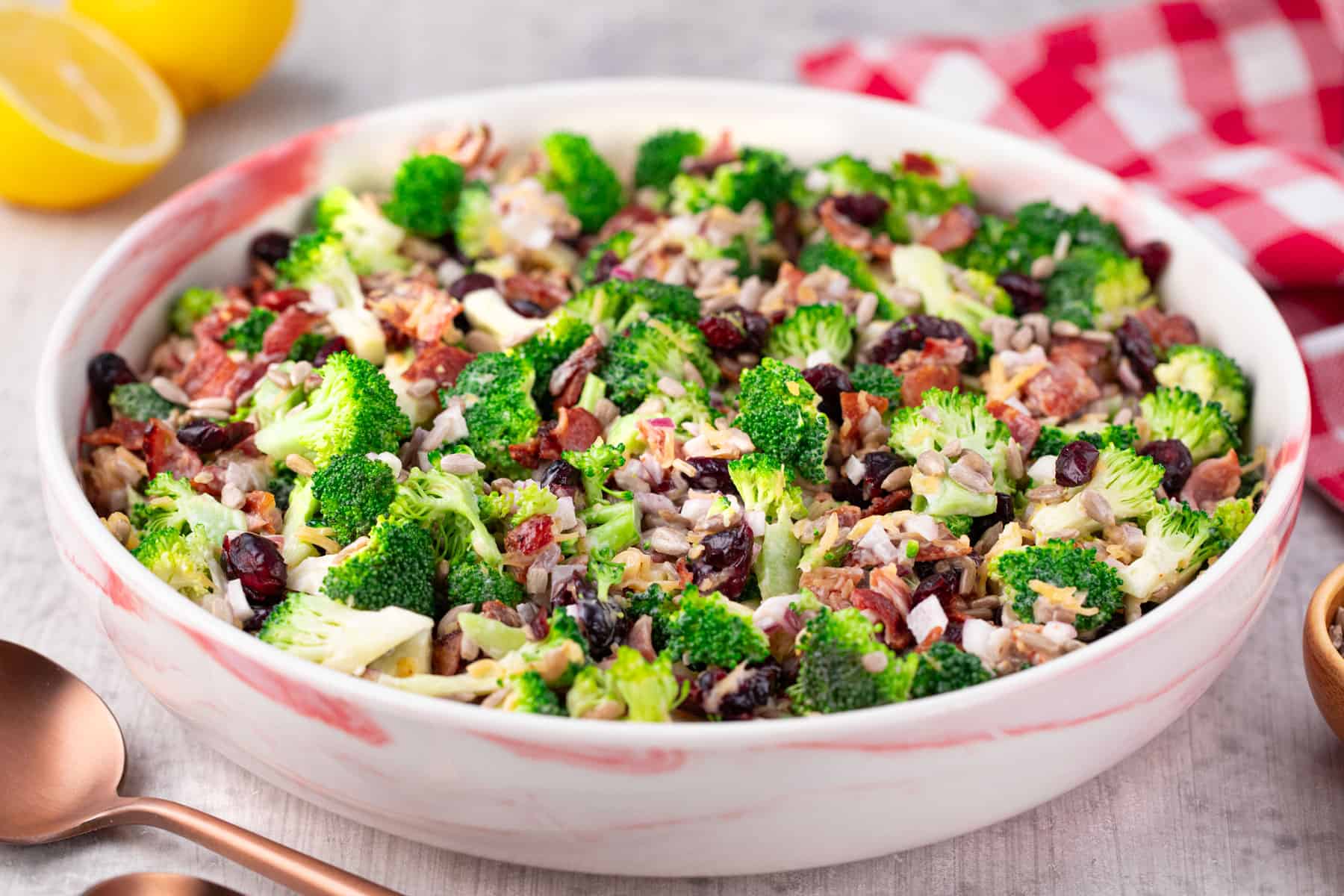 A large white bowl filled with a fresh Costco broccoli salad, featuring chopped broccoli, bacon bits, dried cranberries, sunflower seeds, and a creamy dressing. A red-checked napkin and lemons are in the background.