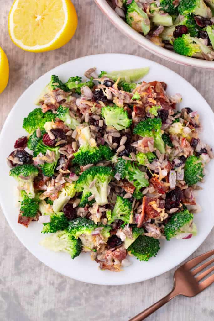 A plate of Costco broccoli salad with sunflower seeds, dried cranberries, red onion, and bacon pieces sits on a table next to a halved lemon and fork. A bowl of extra salad is visible in the background.