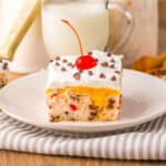 A square slice of cherry chip poke cake topped with whipped cream, mini chocolate chips, and a cherry on a white plate.