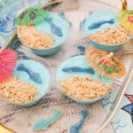 A tray of blue yogurt cups topped with gummy sharks, crushed crackers, and colorful paper umbrellas.