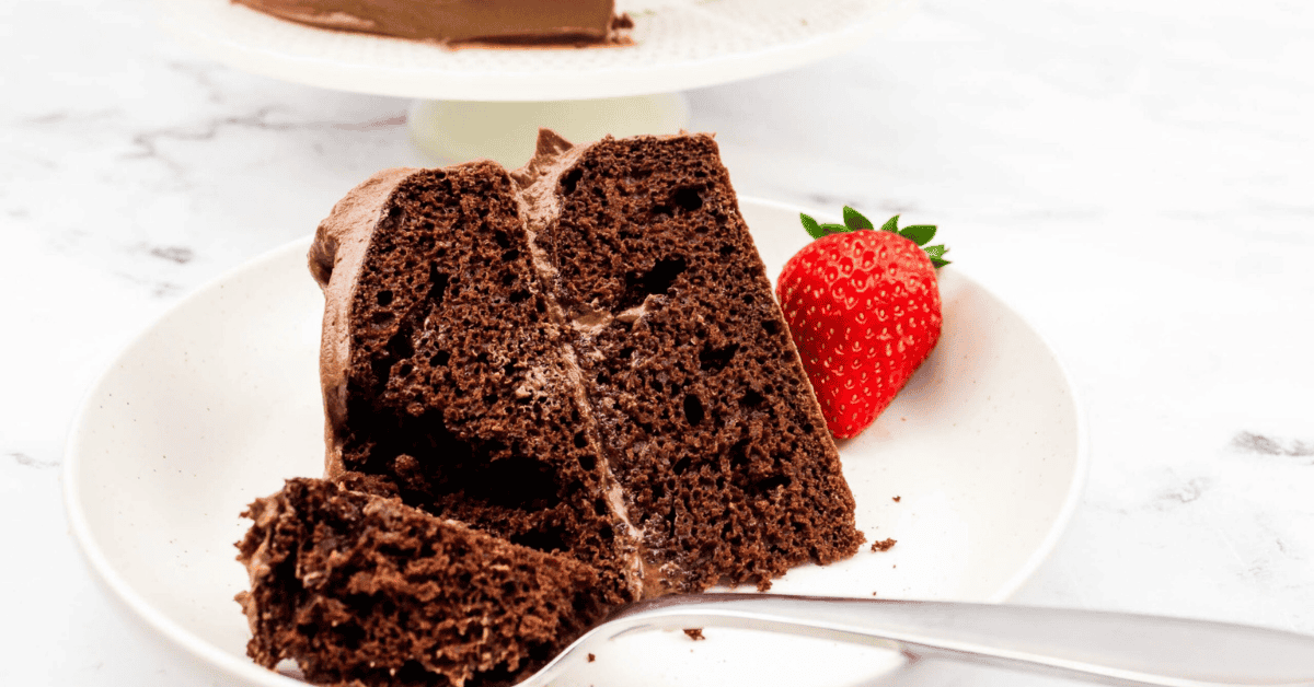 A slice of chocolate cake with chocolate frosting on a white plate, served with a fresh strawberry. The fork suggests you’ve mastered how to make a box cake taste like a bakery cake—a tempting second slice waits in the background.