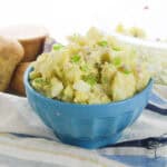 A blue bowl filled with potato salad topped with chopped green onions. Potatoes, a red onion, a salt container, and a pepper grinder are nearby.