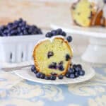 A close-up of a slice of blueberry bundt cake topped with icing and fresh blueberries on a white plate.
