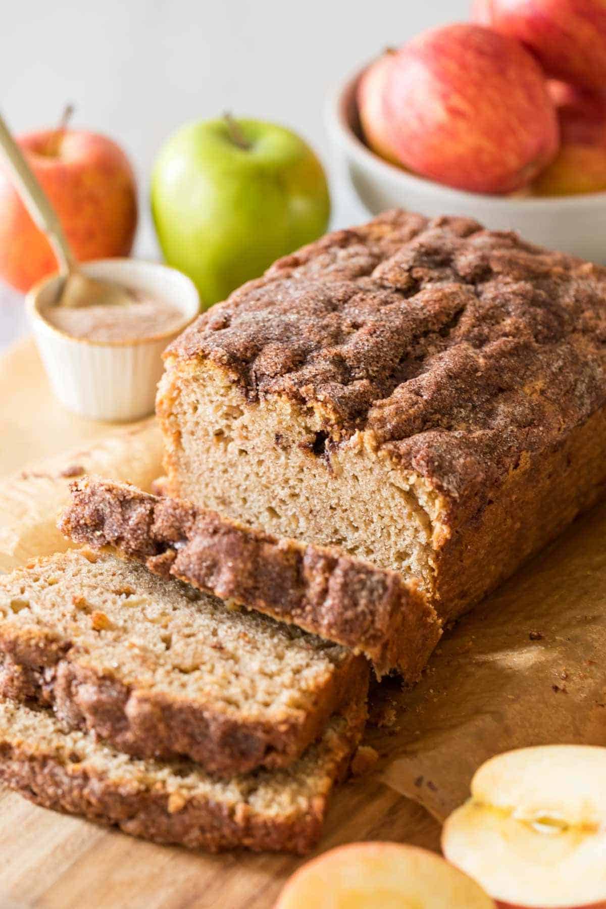 A loaf of cinnamon apple bread sits on a wooden board, partially sliced. Fresh apples and a small bowl of cinnamon sugar are in the background—one of the best easy recipes for leftover apples.