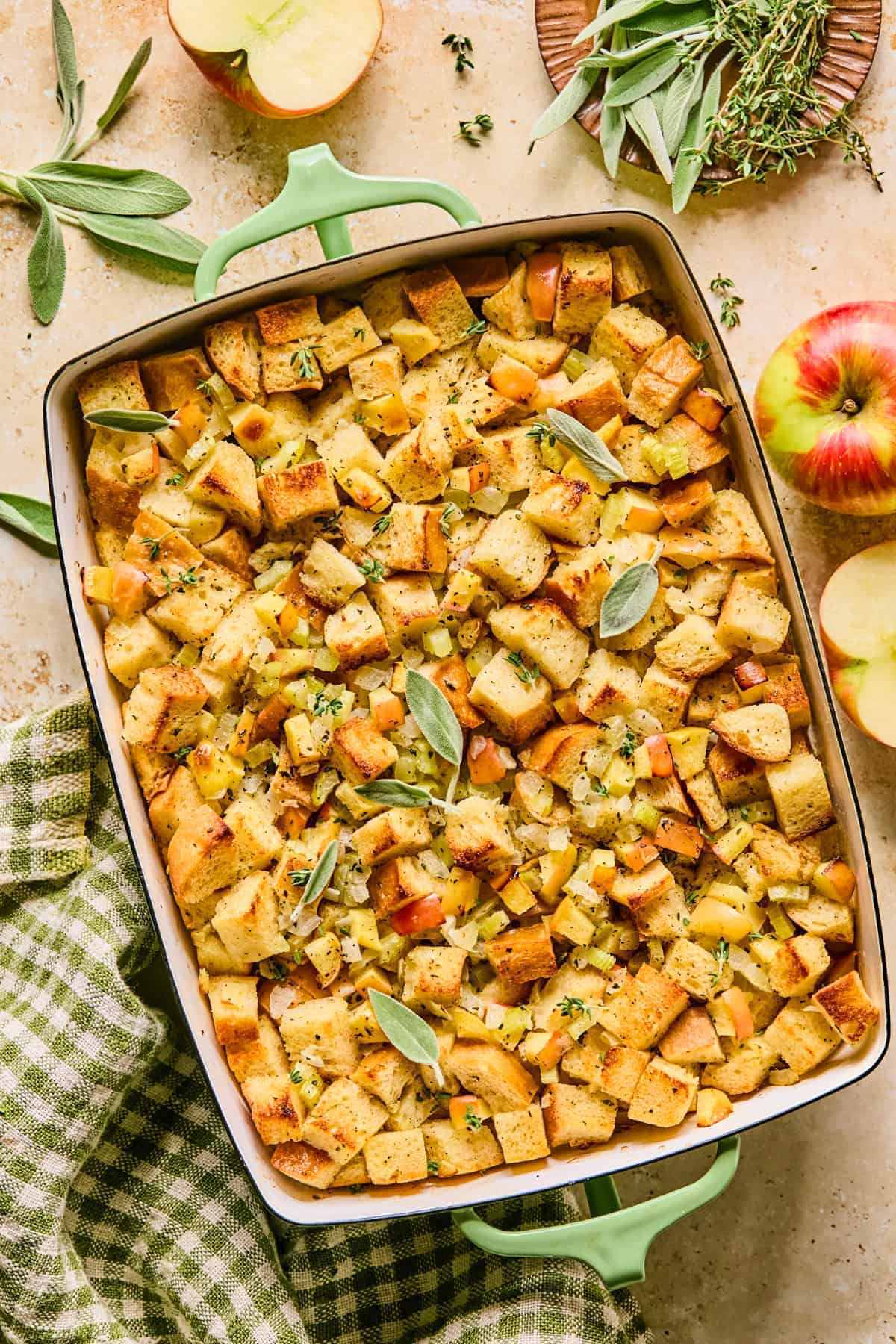 A baking dish filled with golden-brown bread stuffing mixed with apple chunks and herbs, garnished with fresh sage—one of the easy recipes for leftover apples. Nearby are whole apples, fresh herbs, and a green checkered cloth.