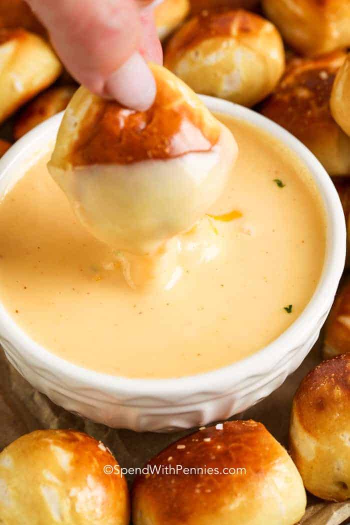 A hand dips a soft pretzel bite into a bowl of creamy cheese sauce, surrounded by more pretzel bites—an irresistible choice for fall party snacks.