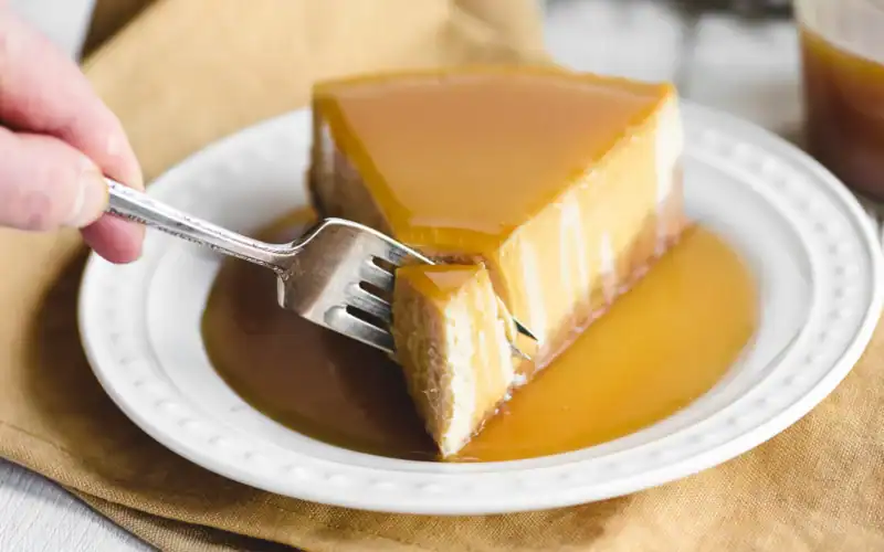 A hand holds a fork, cutting into a slice of cheesecake topped with caramel sauce on a white plate—one of the irresistible fall cheesecake recipes perfect for cozy gatherings.