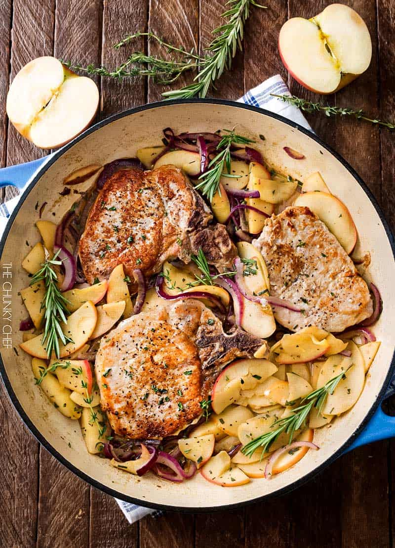 A skillet with three cooked pork chops surrounded by sliced apples, red onions, and rosemary—an inviting dish on a wooden table, perfect for those seeking easy recipes for leftover apples.