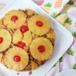 A whole pineapple upside-down cake on a white plate next to a pineapple-patterned napkin and a stack of forks.