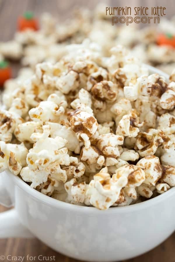 A white mug filled with popcorn topped with pumpkin spice seasoning makes for the perfect fall party snack. Out-of-focus candy pumpkins are in the background. Text reads Pumpkin Spice Latte Popcorn.