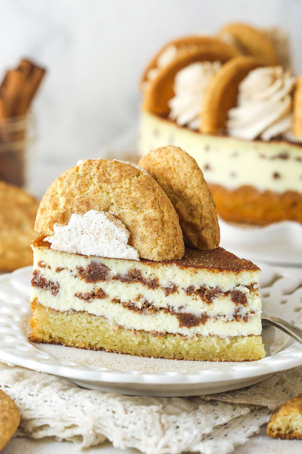 A slice of layered cheesecake with cinnamon cookies and cream sits on a white plate, topped with whipped cream and a whole cookie. Perfect for anyone searching for cozy fall cheesecake recipes, this indulgent dessert is sure to impress. The full cheesecake is in the background.