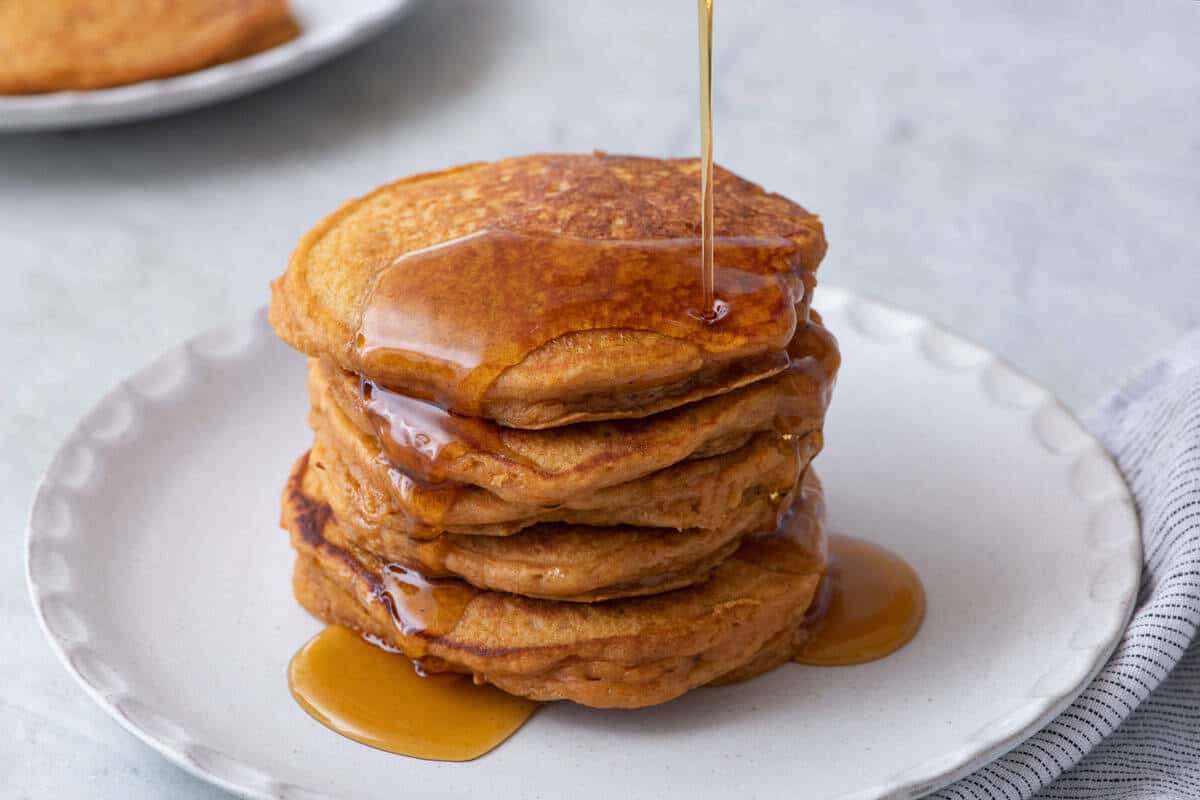 A stack of four pancakes on a white plate with syrup being poured over them—an irresistible scene perfect for cozy mornings and discovering new fall pancake recipes. A striped napkin is to the right, and another plate with pancakes is in the background.