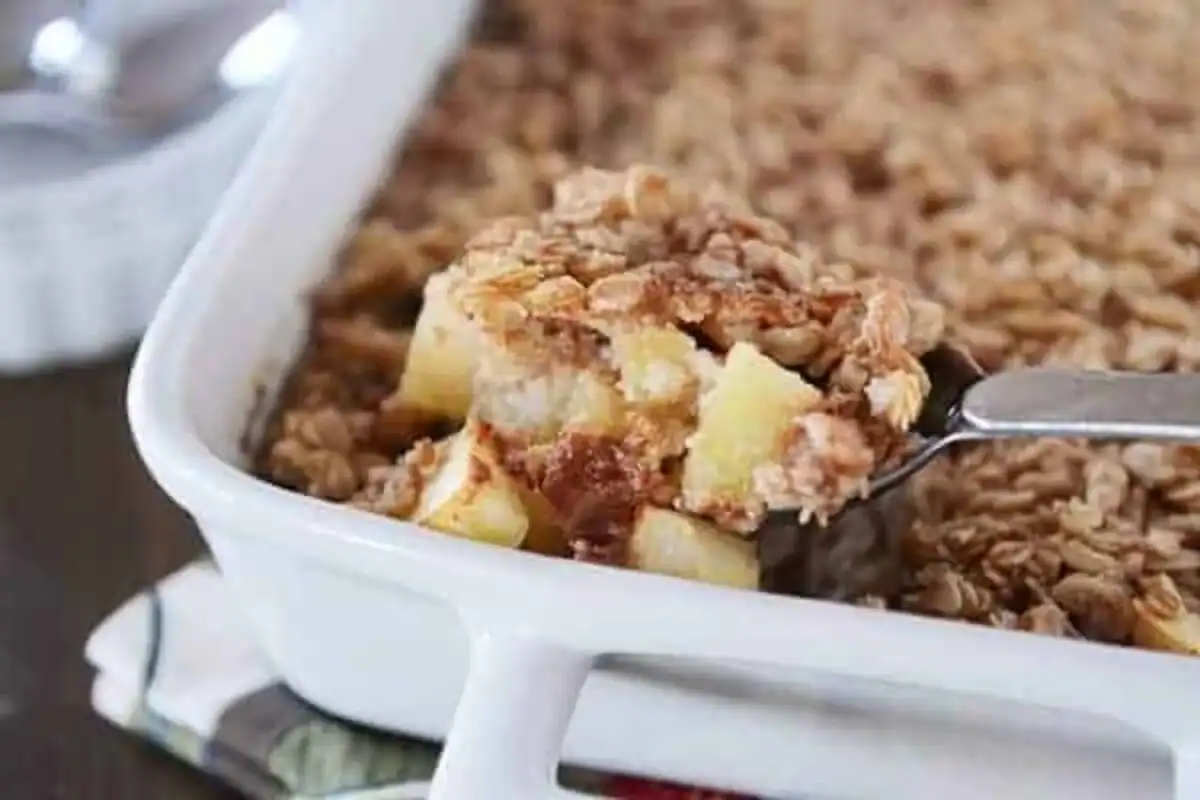 A close-up of a white baking dish filled with apple crisp, one of the coziest fall breakfast recipes. A spoon is lifting a serving, showing chunks of baked apples topped with a golden oatmeal crumble.