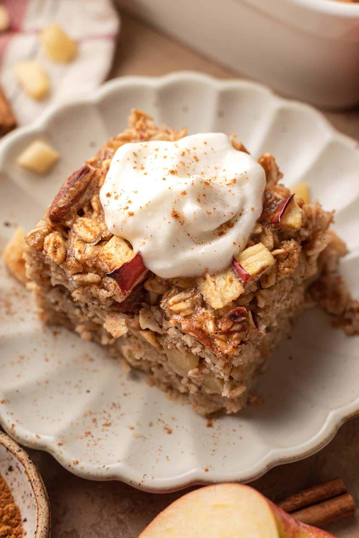 A square piece of baked oatmeal with apple chunks—an ideal way to use up leftover apples—topped with whipped cream and a sprinkle of cinnamon, served on a scalloped plate.