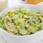 A white bowl filled with a creamy salad containing sliced avocado, zucchini noodles, shredded crab sticks, and topped with sesame seeds. The background shows a jar of grains and a wooden container.