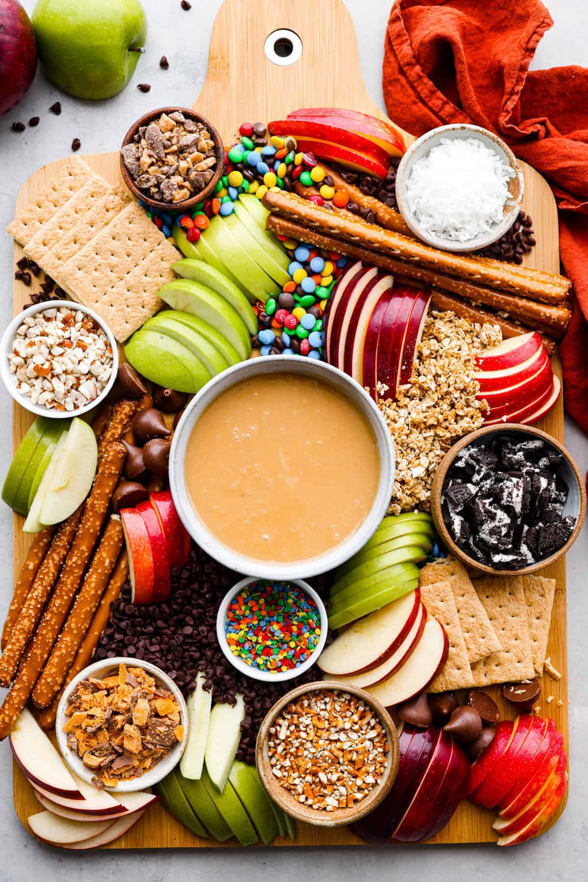 A wooden board with caramel dip in the center, surrounded by sliced apples, graham crackers, pretzel rods, chocolate chips, sprinkles, chopped nuts, candies, shredded coconut, and crushed cookies—perfect for serving as festive fall party snacks.