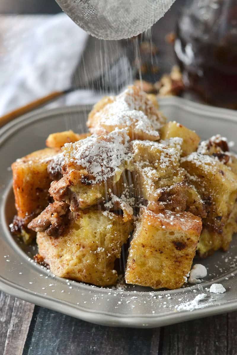 A plate of bread pudding, reminiscent of classic slow cooker apple recipes, is being dusted with powdered sugar, highlighting the chunky, golden pieces. The dessert sits on a decorative plate on a rustic wooden surface.
