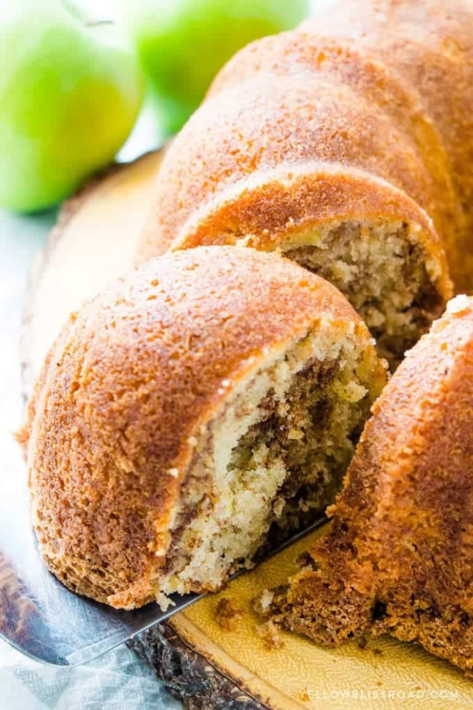 A close-up of a sliced bundt cake with a golden-brown crust and a moist, marbled interior, displayed on a wooden board with green pears in the background—an inviting centerpiece that’s perfect for your fall cake ideas.