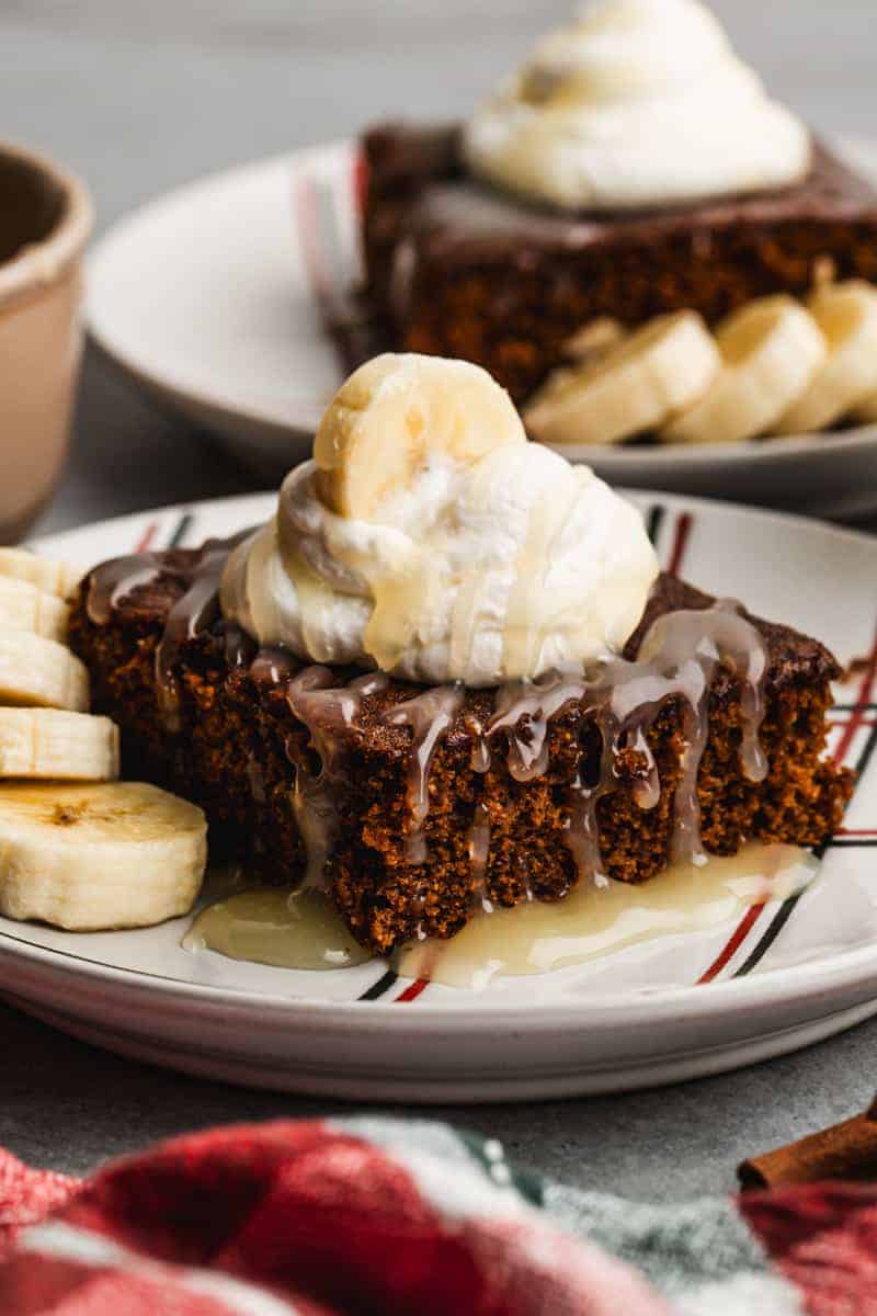 A slice of gingerbread cake topped with whipped cream, banana slices, and sauce, served on a plate with extra banana slices on the side—this delicious treat is perfect for anyone searching for fall cake ideas.
