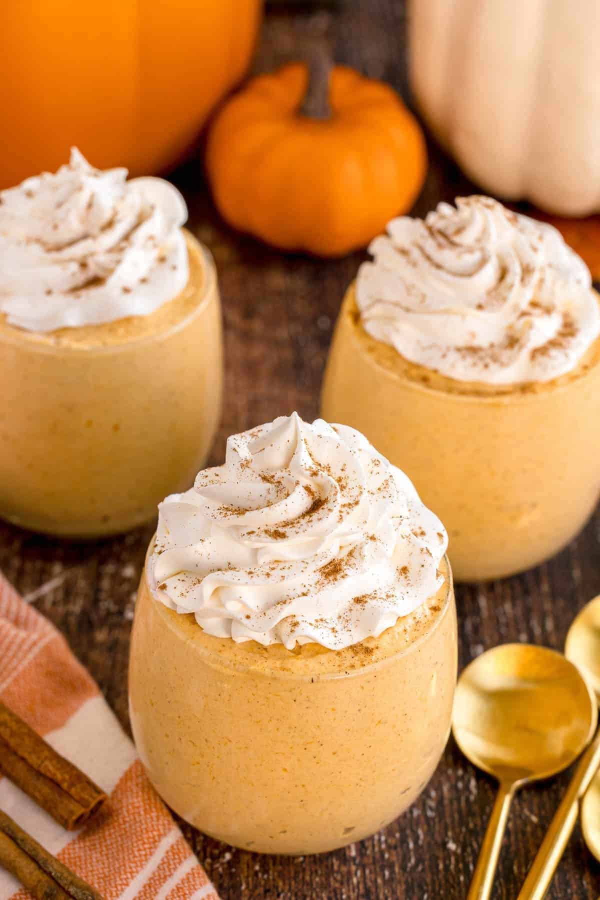 Three glasses filled with creamy pumpkin mousse—one of our favorite mini pumpkin dessert recipes—are topped with whipped cream and sprinkled cinnamon, set on a wooden table with gold spoons, cinnamon sticks, and small pumpkins in the background.
