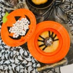 A glazed donut decorated as a spider with licorice legs and a chocolate head on an orange plate, next to a pumpkin-shaped dish filled with small white candies, all on a spooky, Halloween-themed table setting.