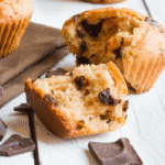 A Bisquick chocolate chip muffin broken in half sits on a white wooden surface, surrounded by pieces of chocolate and another whole muffin on a brown napkin in the background.