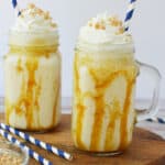 Two mason jar glasses of caramel milkshake, inspired by a Butterbeer Frappuccino Recipe, are topped with whipped cream, chopped nuts, and blue-striped straws. Caramel sauce drips down the sides, with extra nuts in a small dish nearby.
