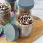 Mason jars filled with chocolate cake and topped with swirls of chocolate frosting and colorful sprinkles sit on a wooden board, showcasing this delightful Chocolate Cake in a Jar Recipe. A multicolored cloth is partially visible in the background.