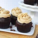 Four chocolate cupcakes with swirls of coffee cream cheese frosting are arranged on a white rectangular plate. Coffee beans are scattered nearby, and more cupcakes are displayed on a white cake stand in the background.