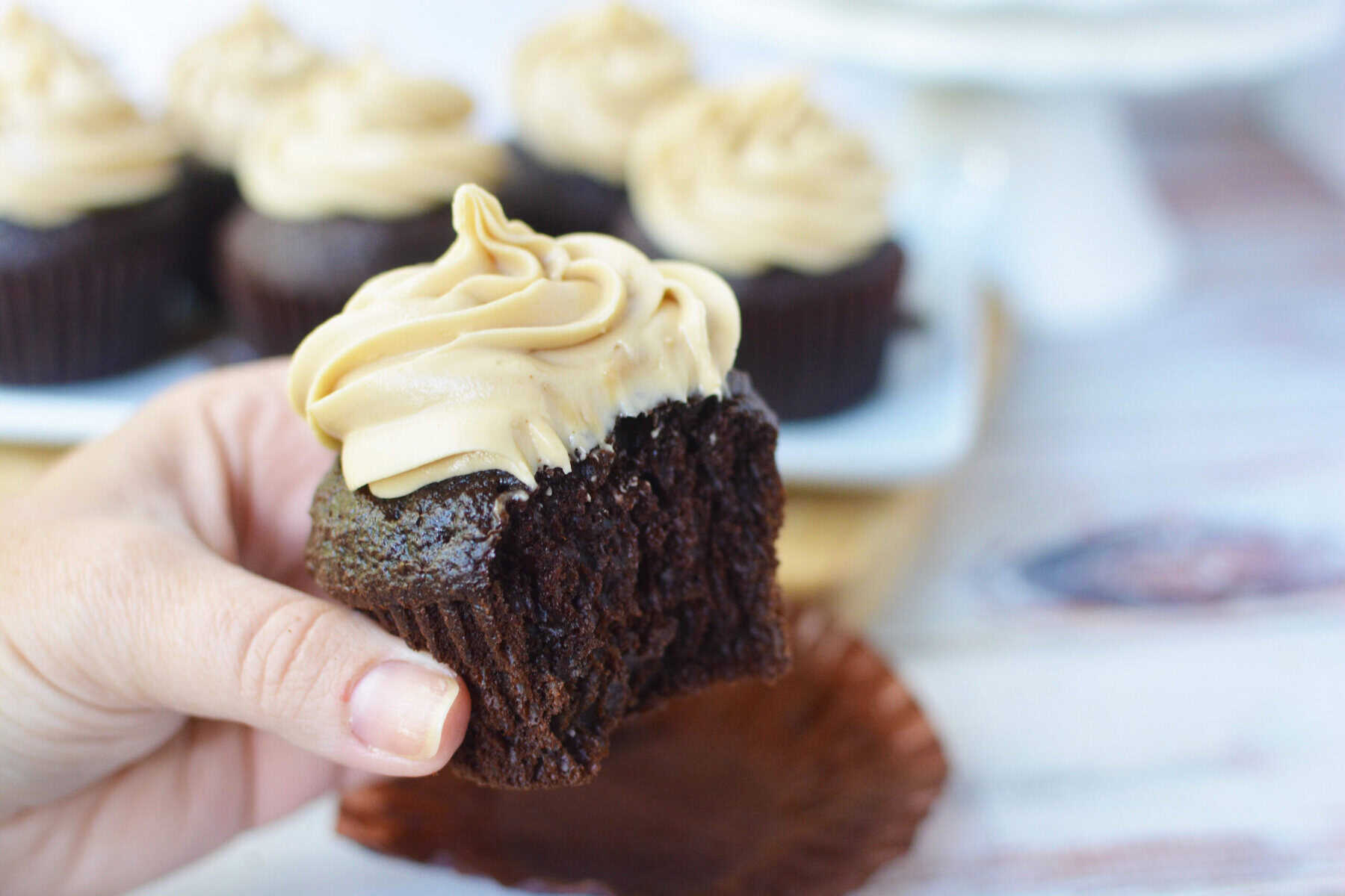 A hand holding a chocolate cupcake with coffee cream cheese frosting and a bite taken out, with more cupcakes topped with the same rich frosting in the background on a white tray.