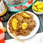 A plate of shredded beef topped with whole pepperoncini peppers, next to a gold fork, a checkered red napkin, salt and pepper containers, a bowl of extra peppers, and a floral-patterned pressure cooker.