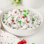 A white bowl filled with Grinch Puppy Chow, coated in powdered sugar and topped with red and green candy pieces, sits on a white surface next to a white napkin with festive red and green dots.