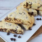 A close-up of Bisquick Chocolate Chip Scones arranged on a white rectangular plate, with a few chocolate chips scattered around. The scones are golden brown with visible chocolate chips, set against a rustic wooden surface.
