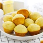 A white plate filled with golden cornbread muffins sits on a white surface, surrounded by an egg, a glass jar of cornmeal, and a checkered kitchen towel—perfect for homemade cornbread muffins lovers.