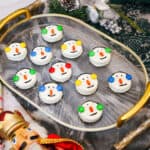 A tray of round cookies decorated as snowman faces with candy eyes, orange carrot-shaped noses, and colorful ear muffs, surrounded by festive holiday decorations.