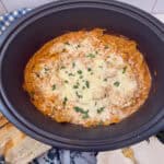 A slow cooker filled with cheesy baked lasagna in a creamy pink sauce, topped with melted cheese and chopped herbs, sits on a blue checkered cloth next to sliced bread and a plate with a fork.