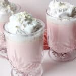 Three clear glass mugs filled with light pink hot chocolate, topped with whipped cream and white chocolate shavings, are arranged on a pale pink surface with a pink cloth in the background.