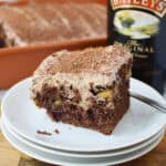 A slice of Bailey’s Poke Cake with creamy frosting and chocolate shavings sits on stacked white plates. In the background, a baking dish with more cake and a bottle of Baileys Irish Cream complete the scene.
