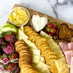 A Valentine’s Day Charcuterie Board featuring assorted crackers, sliced cheeses, ham, grapes, cherry tomatoes, strawberries, pineapple chunks, cashews, and two small bowls of dips is arranged neatly on a marble surface.